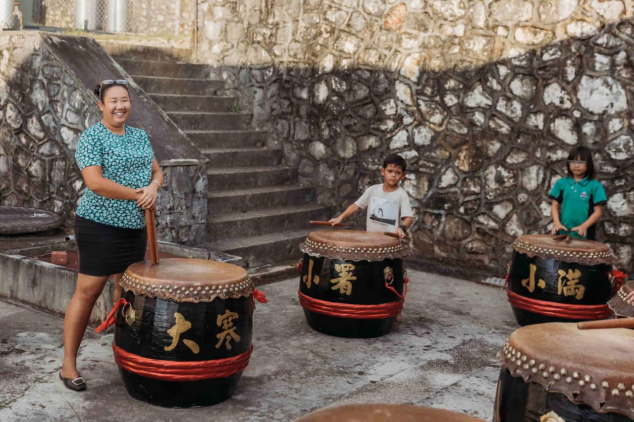 A woman standing next to a large drum on the ground. A boy and a girl are near her and also standing behind large drums