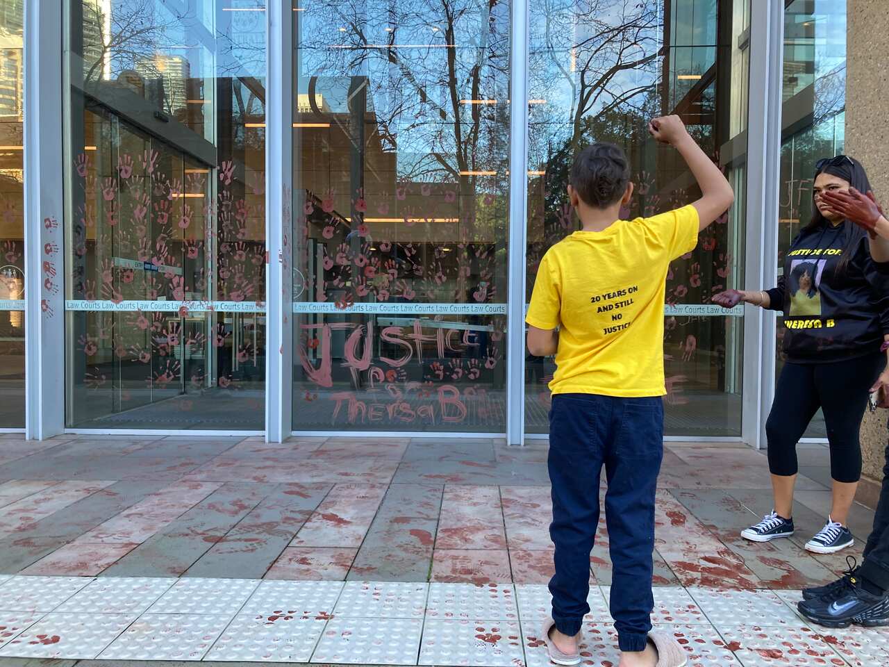 A young boy faces aware from the camera with his fist raised in a black power salute. On the glass wall in front of him ochre hand prints are printed all over. 