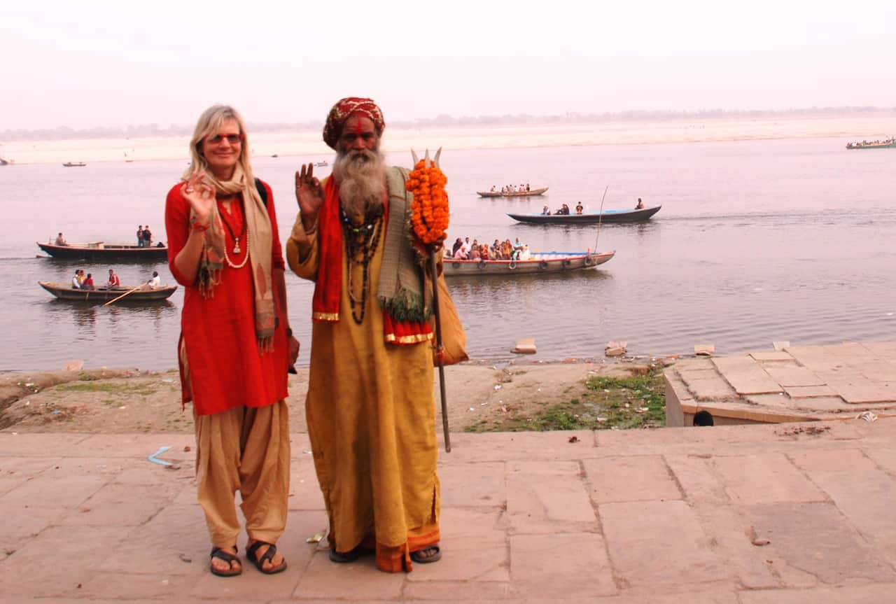 A woman and and Indian man,  both dressed in orange, stand on the bank of a river. There are long narrow boats with people in them in the water behind them. 