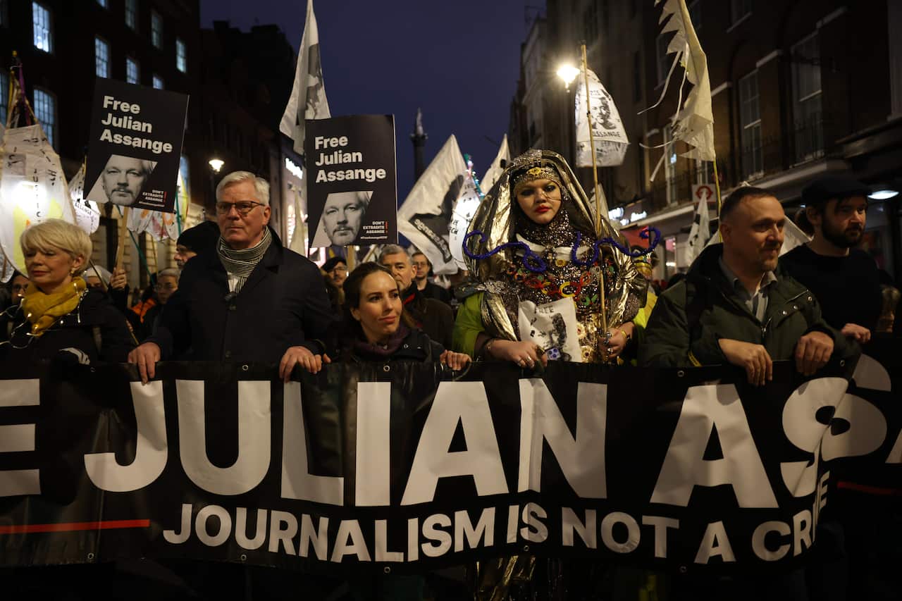 People walking behind a black and white banner which reads in large letters: Free Julian Assange, and below it Journalism is not a crime. Some people are holding posters reading Free Julian Assange