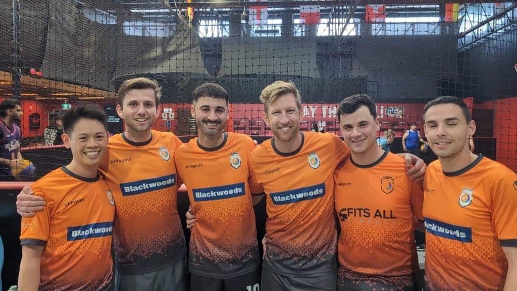 Six men wearing matching orange football jerseys are smiling while embracing each other around the shoulders with an indoor football net in the background