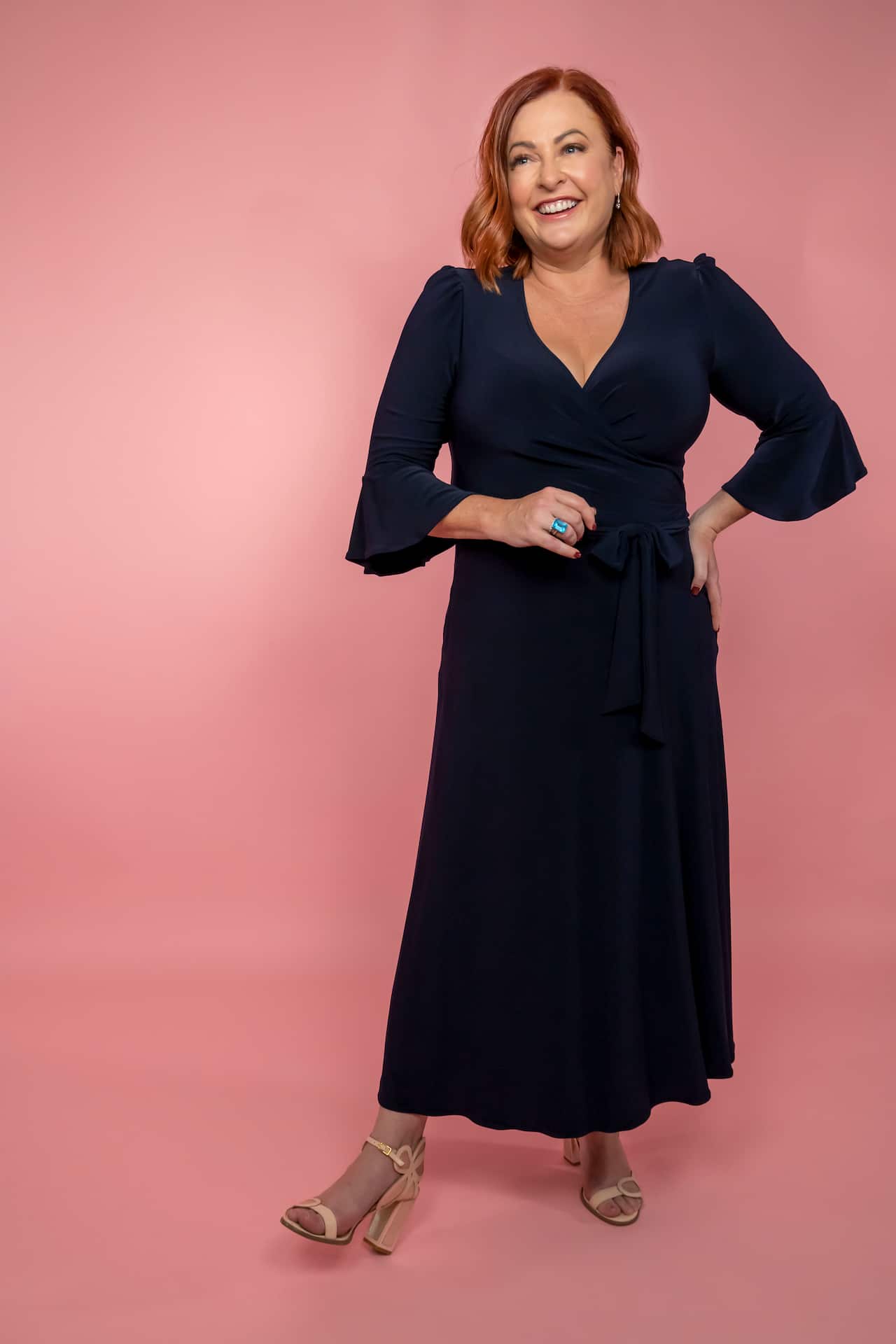 A woman in a black dress stands with one hand on her hip and smiles at the camera against a pink background.