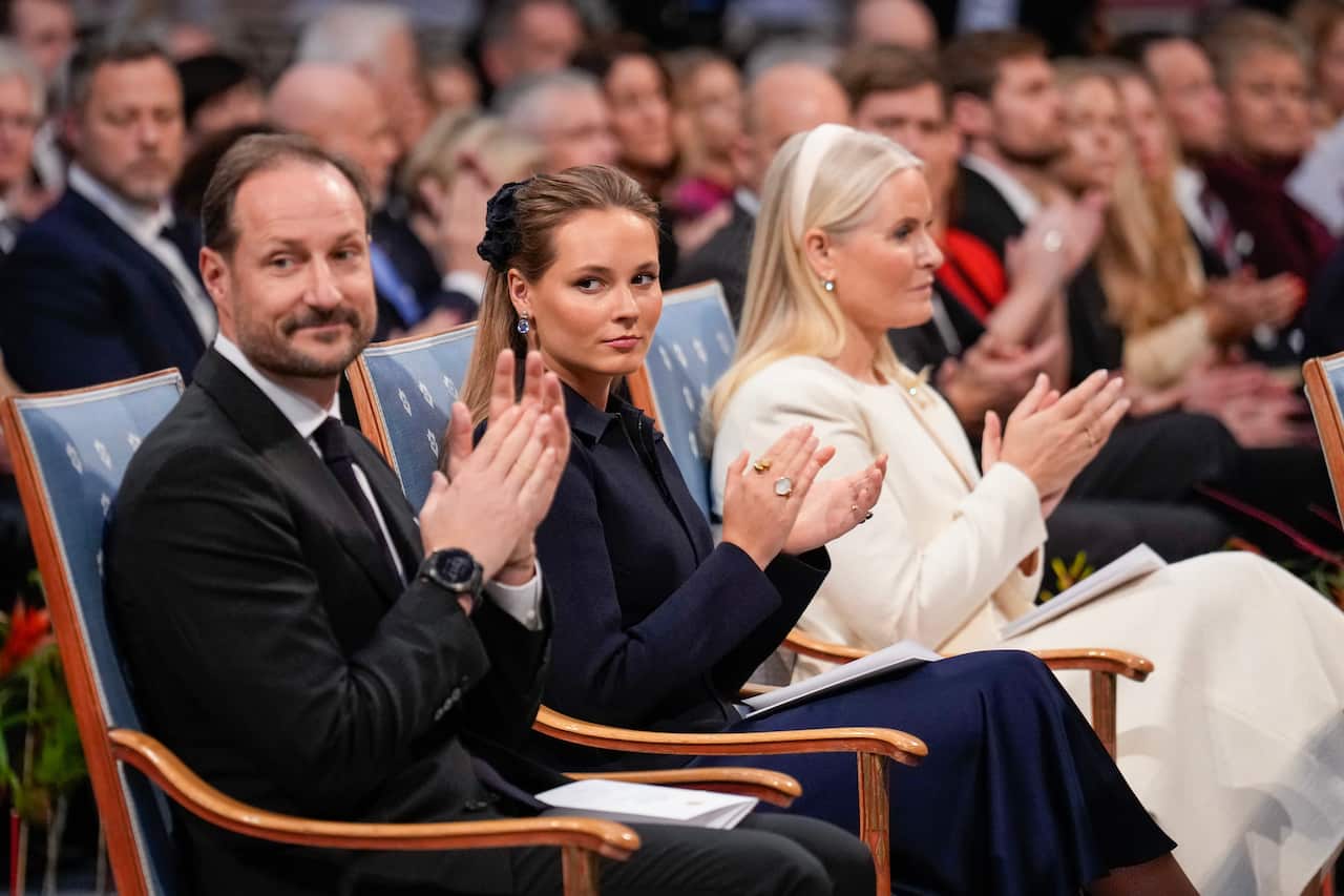 Three people sit in a crowded room and applaud. A young woman seated in the centre has her gaze turned towards one side.