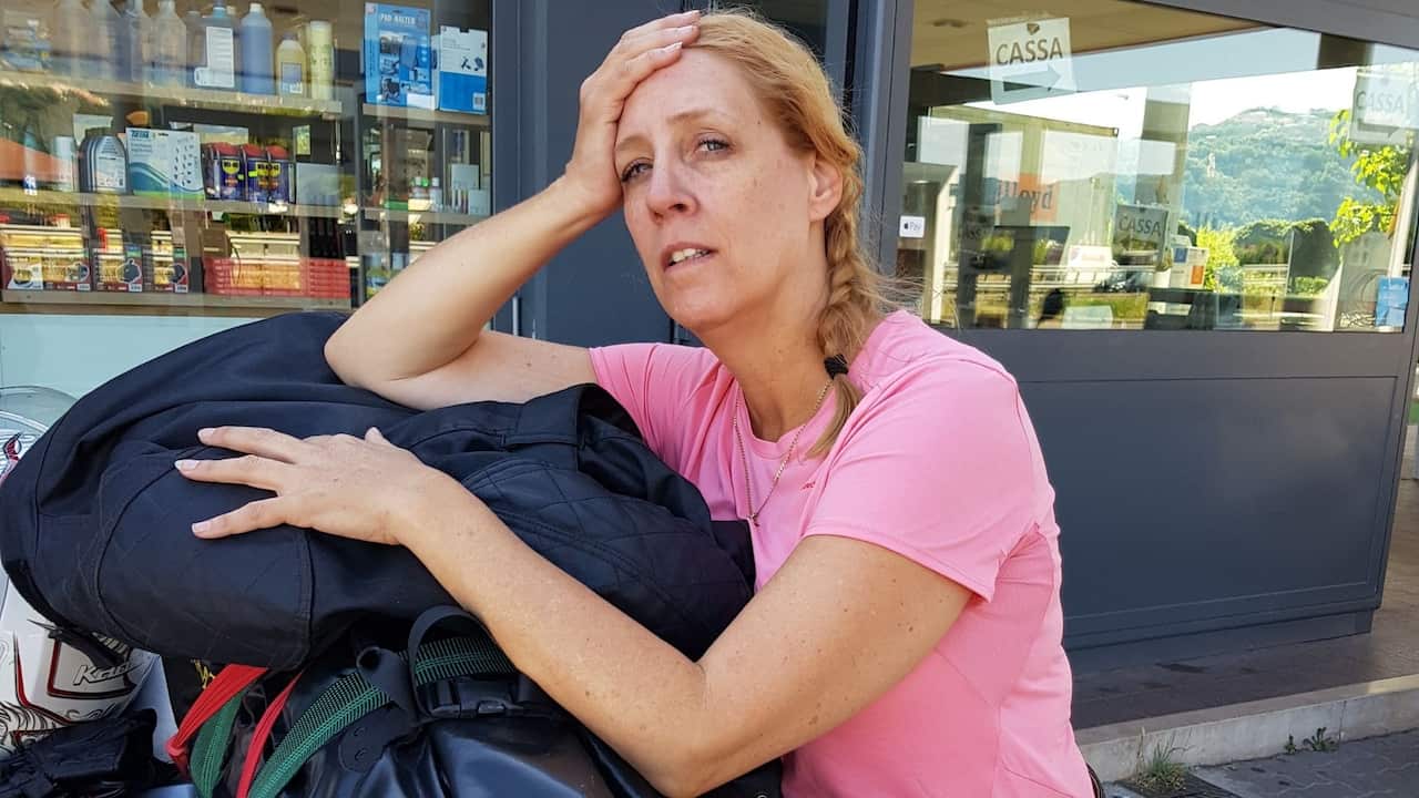 A middle-aged woman with a blonde plait and in a pink t-shirt leans tiredly on a motorbike in front of a shop.