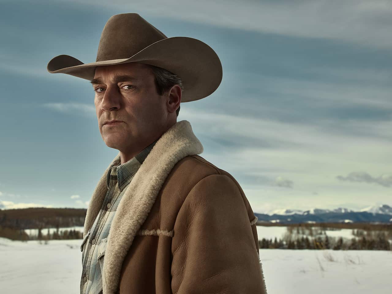 Jon Hamm stands in front of a snowy landscape, looking sideways at the camera. He is wearing a Western hat and fur-collared coat.