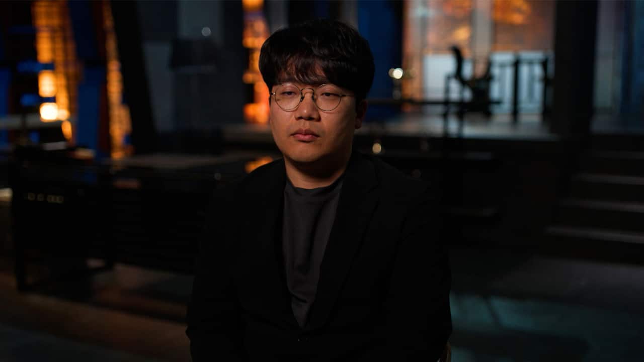 An Asian man wearing round-framed prescription glasses, sitting down for an interview in a TV studio