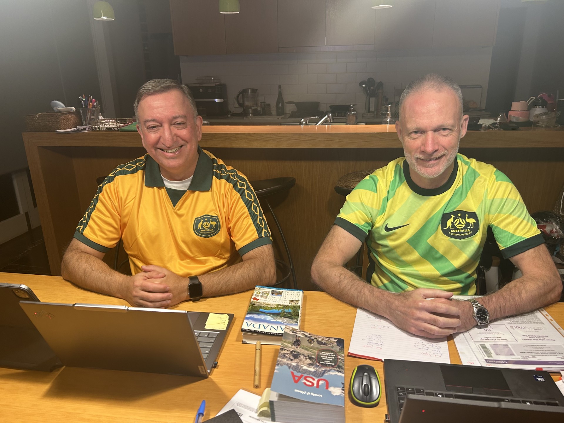 Two middle-aged white men wearing Socceroos jerseys sitting at a desk 