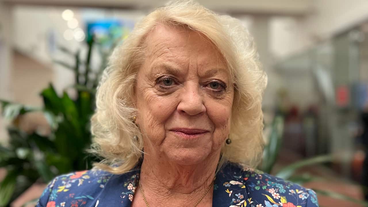 a close-up photo of an older woman with blonde hair in a floral dress in an office