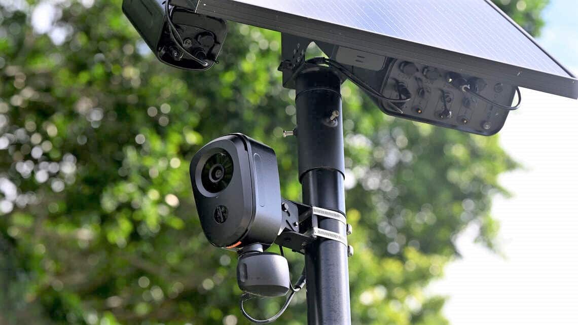 A black Flock safety camera on a pole against a leafy backdrop.
