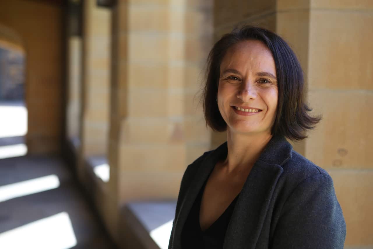 A woman in a blue jacket stands against a sandstone wall.