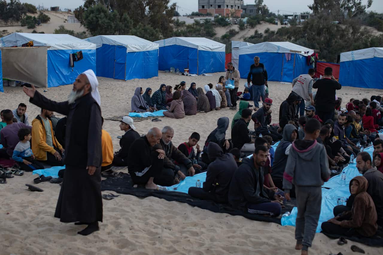 Groups of people sitting down to eat in rows outside. There are blue-and-white tents in the background. A man in the foreground wearing a black gown and white headdress is holding out his right arm in prayer.