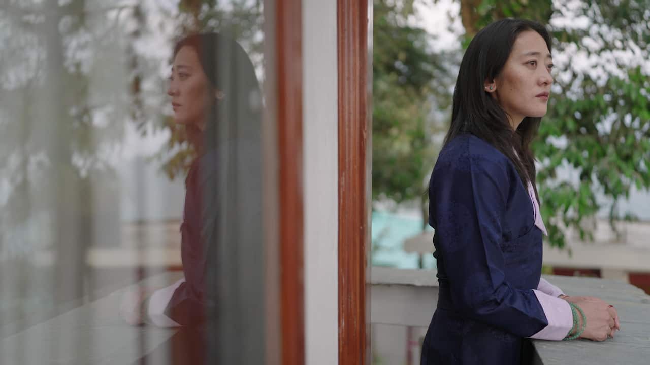 A woman wearing a dark blue outfit and with long dark hair stands on a balcony looking out into the distance. Her reflection can be seen in the window behind.