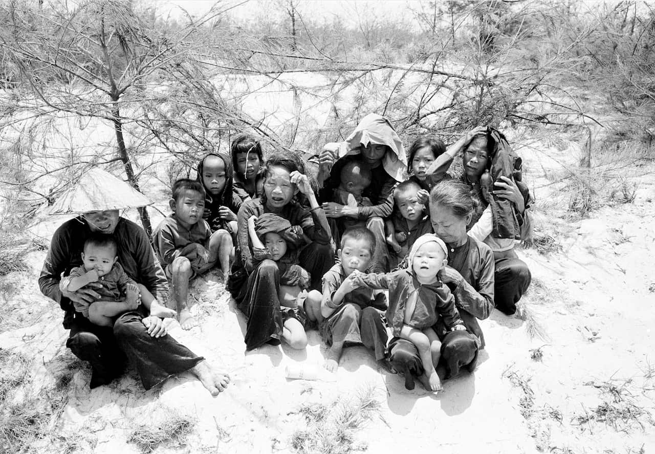 A small group of women and children sitting together on sandy ground near some shrubs