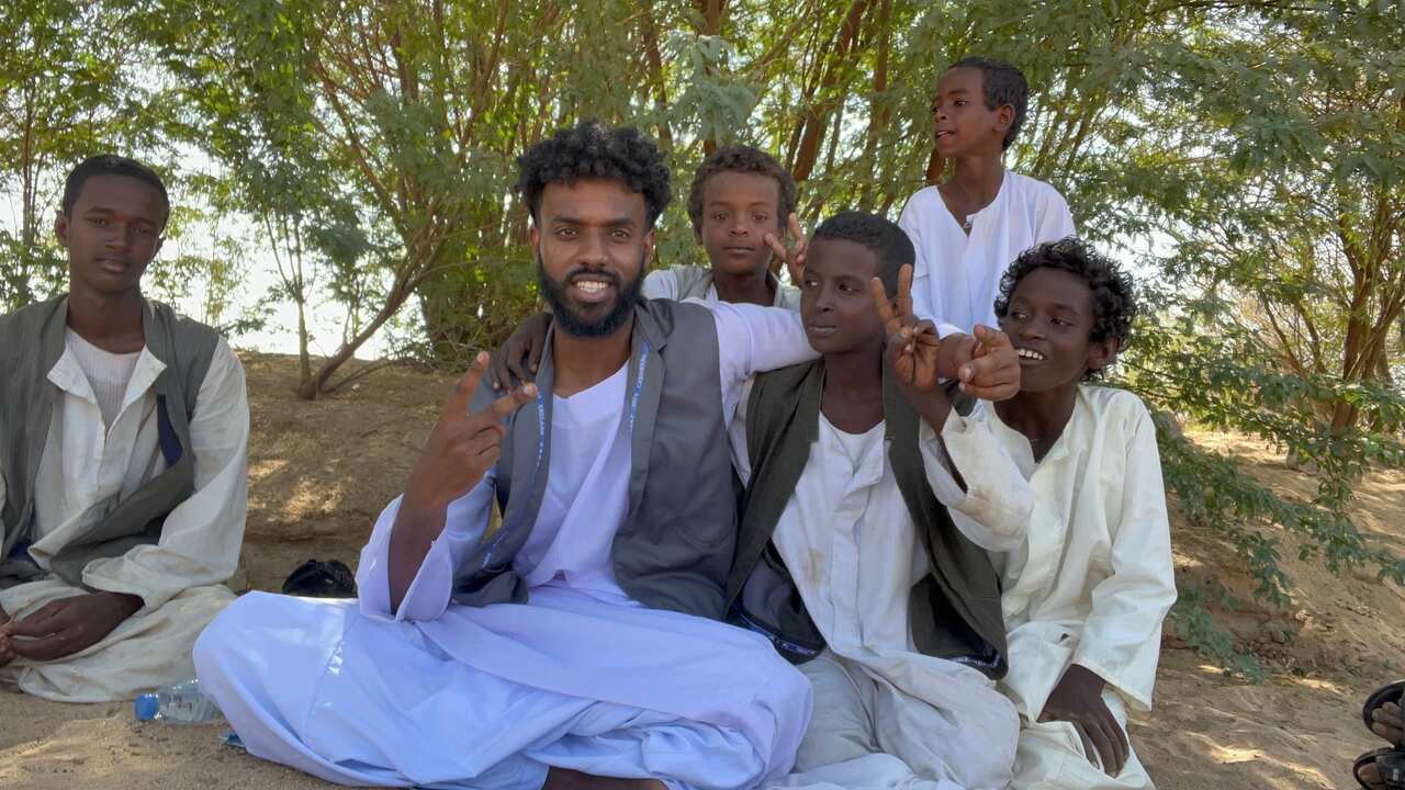 A young man sitting outside on the ground with his legs, holding his fingers up in a peace sign and surrounded by adolescent boys, some also holding up peace signs.