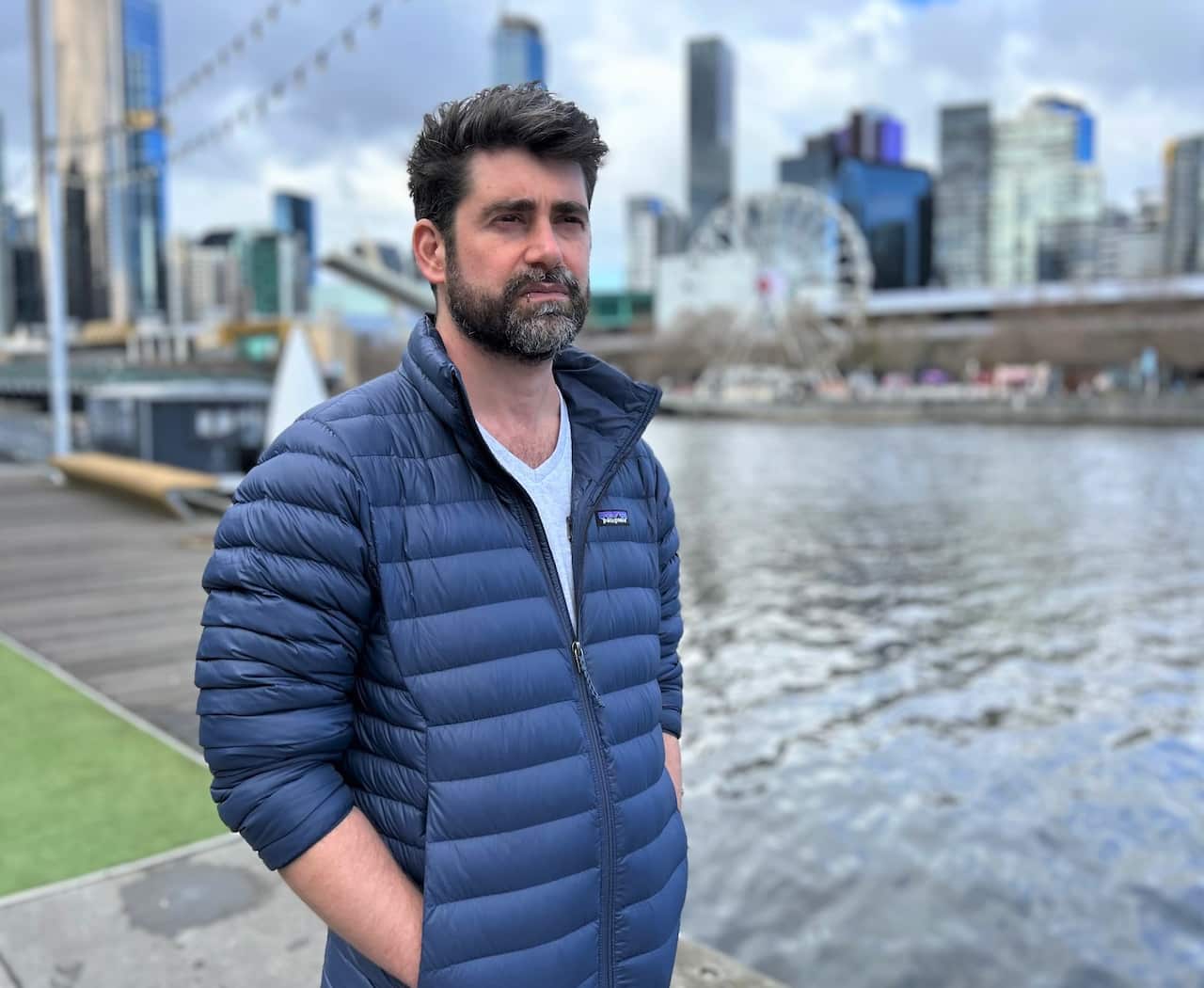 A bearded man in a blue parka stands next to a river with a city backdrop.