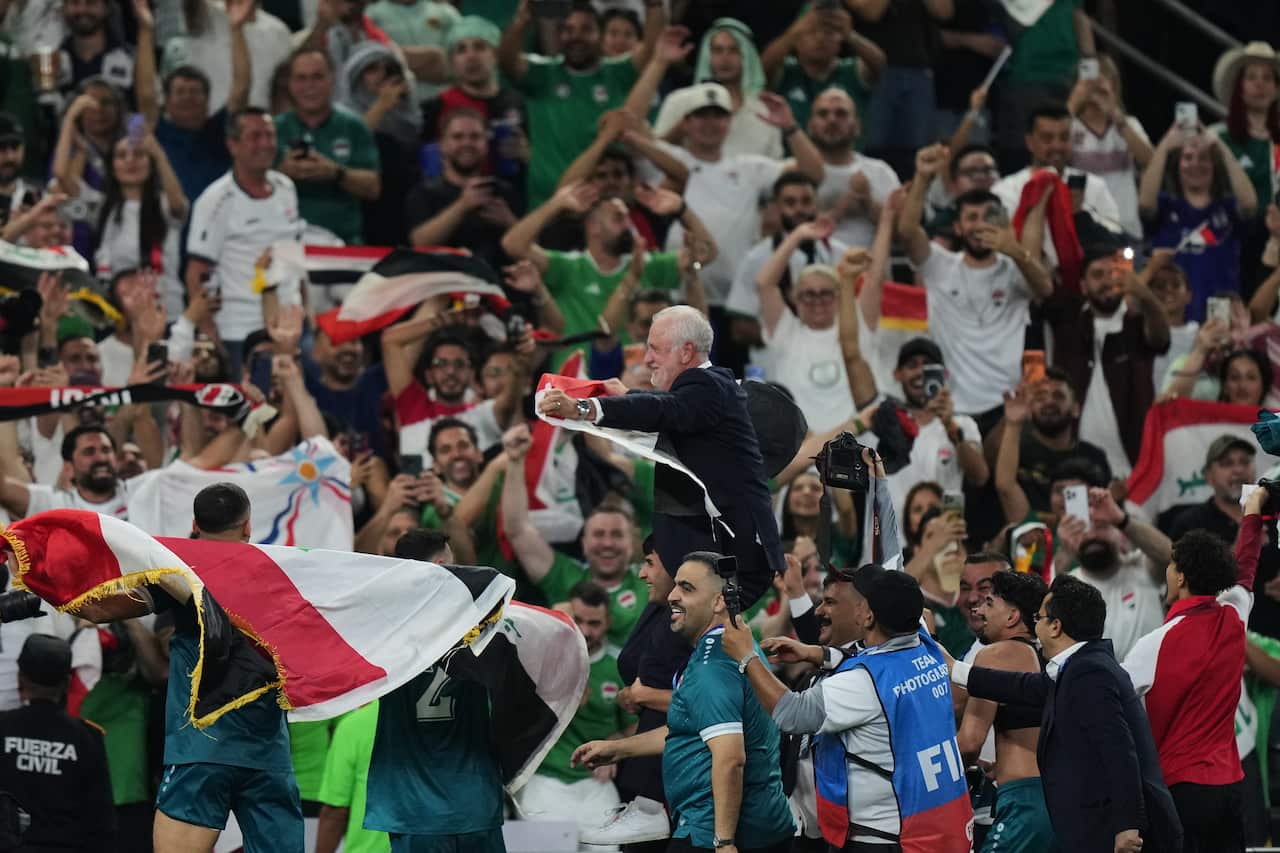 A joyous crowd of fans and players celebrate in a stadium as they hoist a smiling man in a suit onto their shoulders while waving the Iraqi flag.
