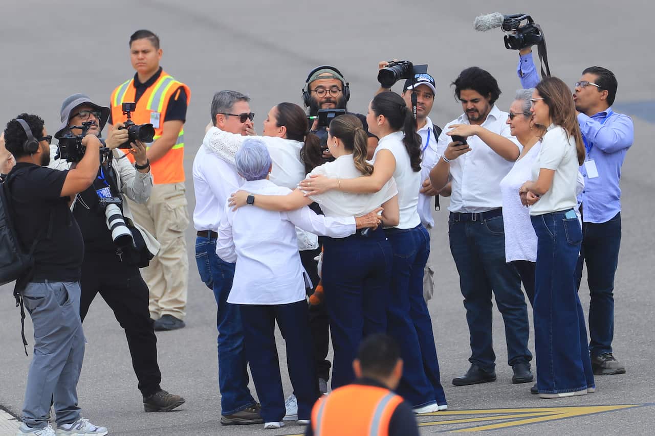 Former Honduran president Juan Orlando Hernández, in a white shirt and blue jeans, hugs his family members on a tarmac, surrounded by media personnel.