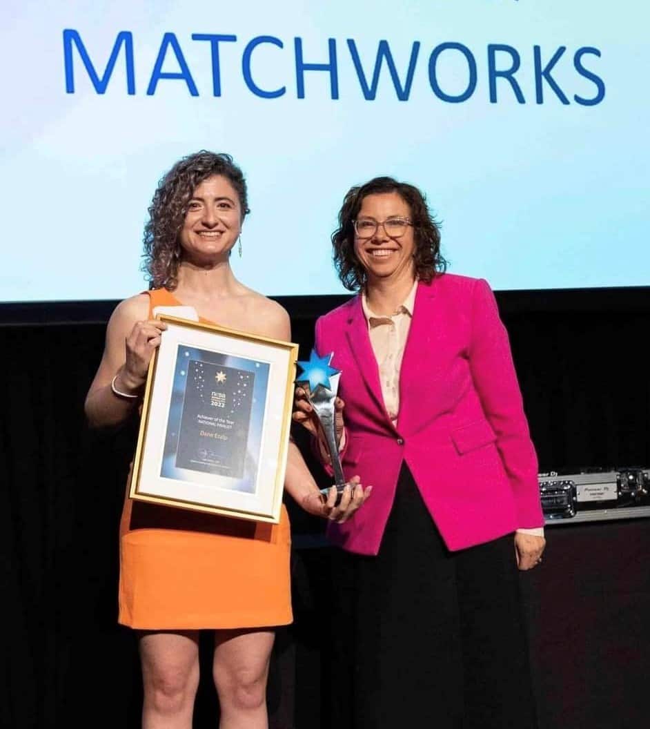 A woman holds a framed certificate and trophy next to another woman. 