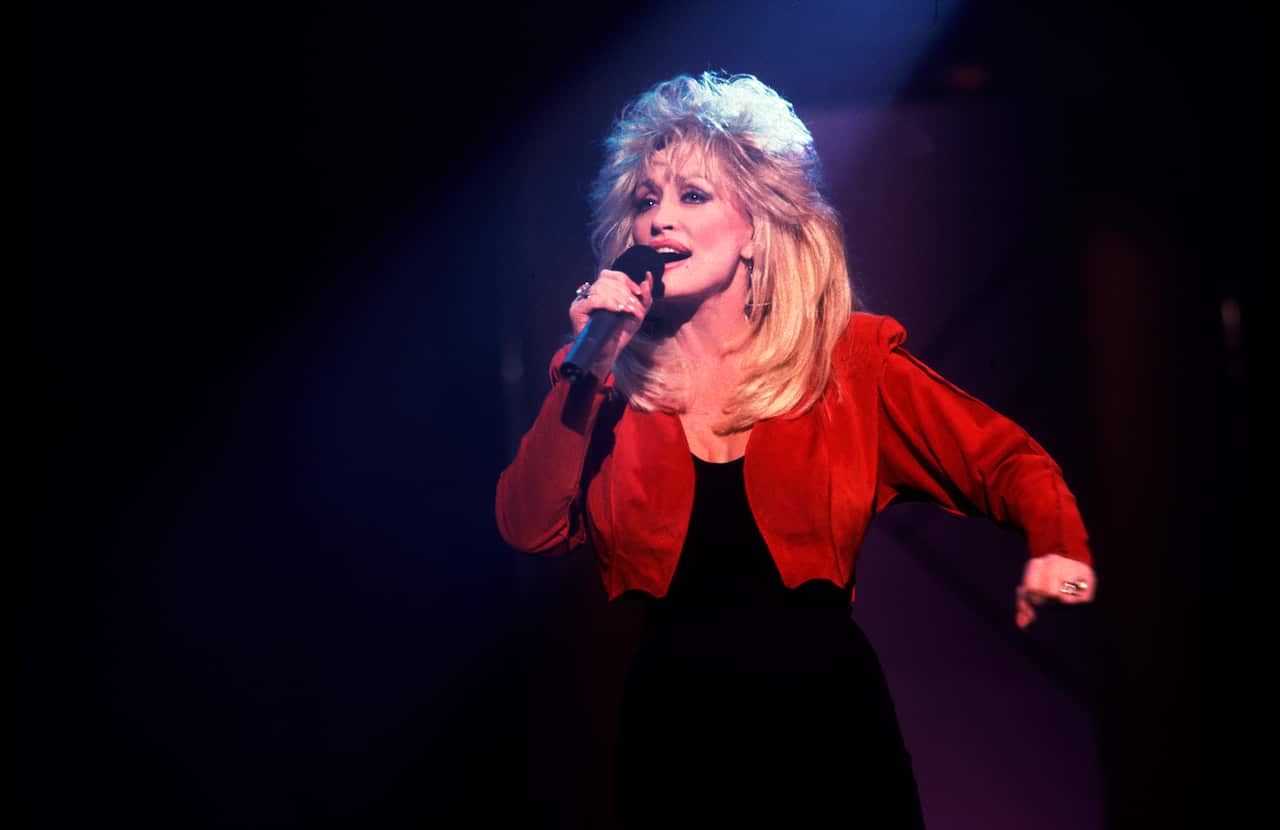 Dolly Parton holding a handheld microphone on stage under dramatic blue and purple lighting, wearing a red jacket over a dark outfit during a live performance.