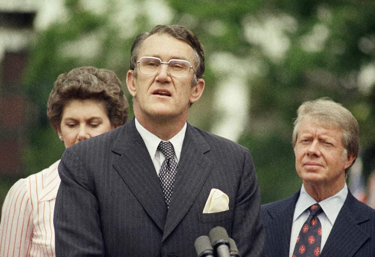 A man in a suit speaking at a microphone. A woman with brown hair and an older man is behind him.