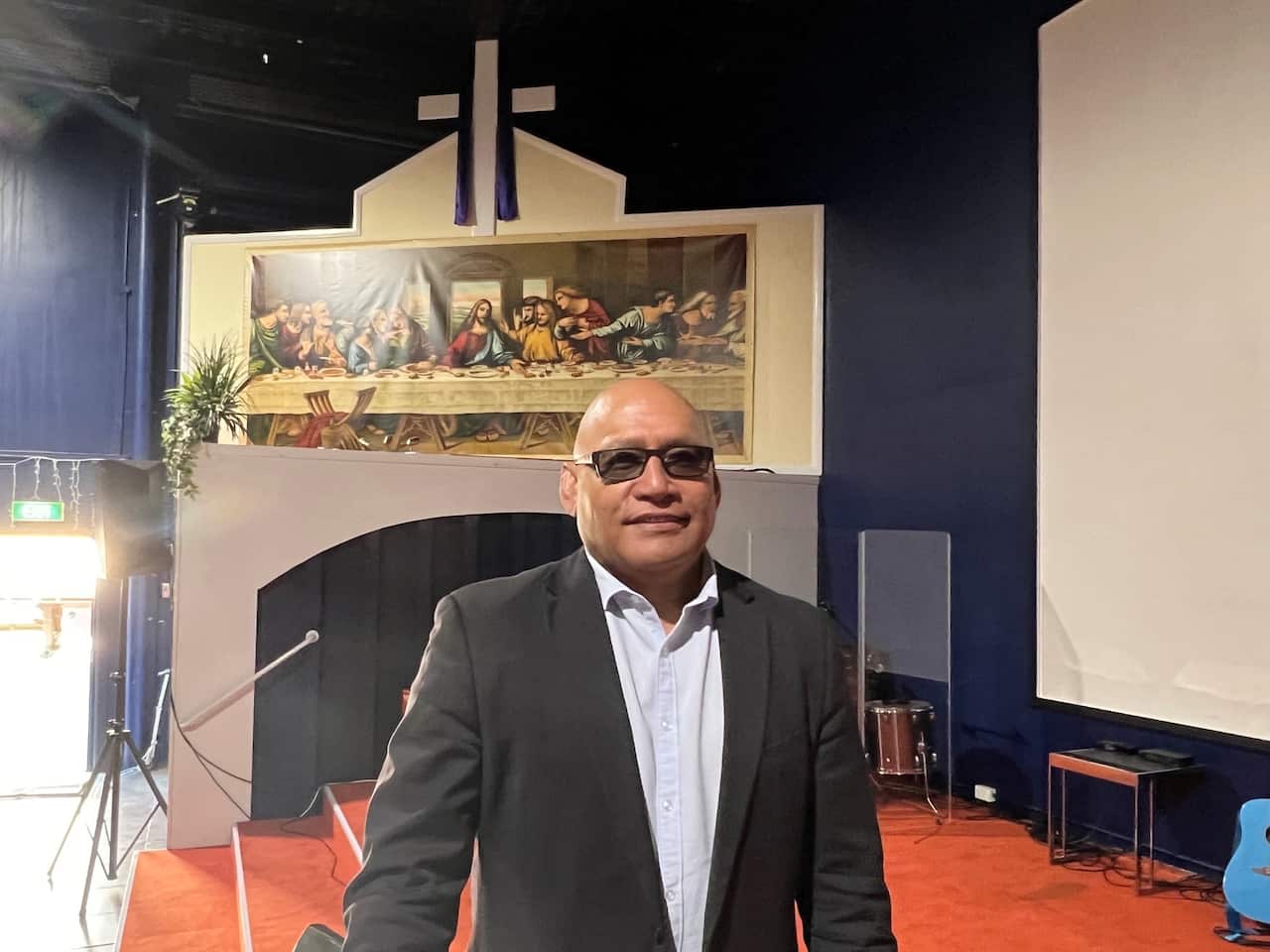 A man in a suit stands in front of a poster of The Last Supper and a cross. 