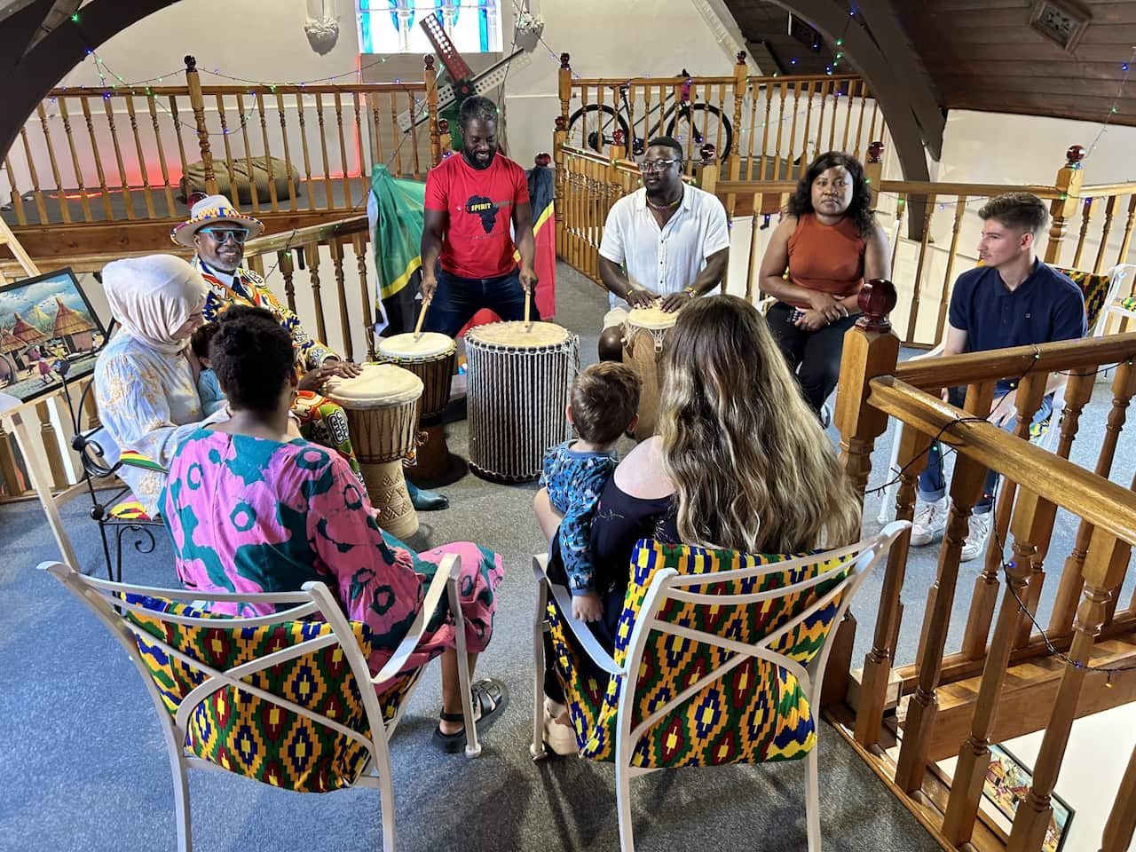 A group of people sitting inside. There are some drums on the floor