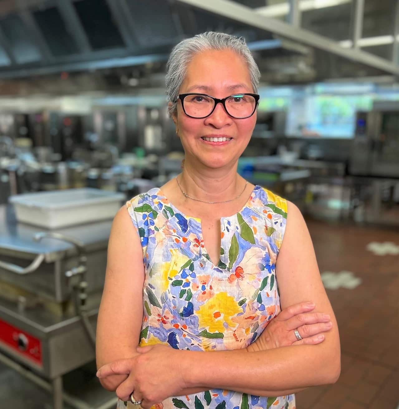 A woman in a floral dress and glasses standing in a commercial kitchen with her arms folded.