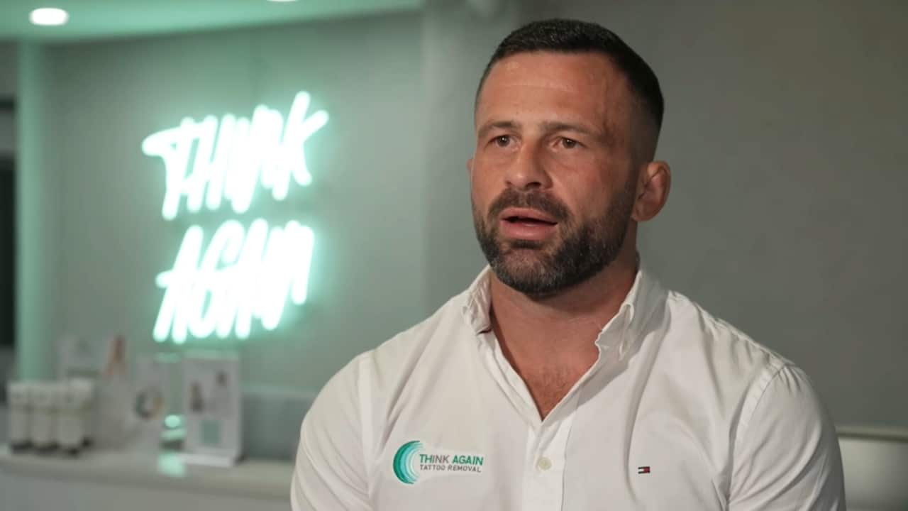 A man in a white shirt talks with a neon sign 'Think Again' plastered on a wall in the background.