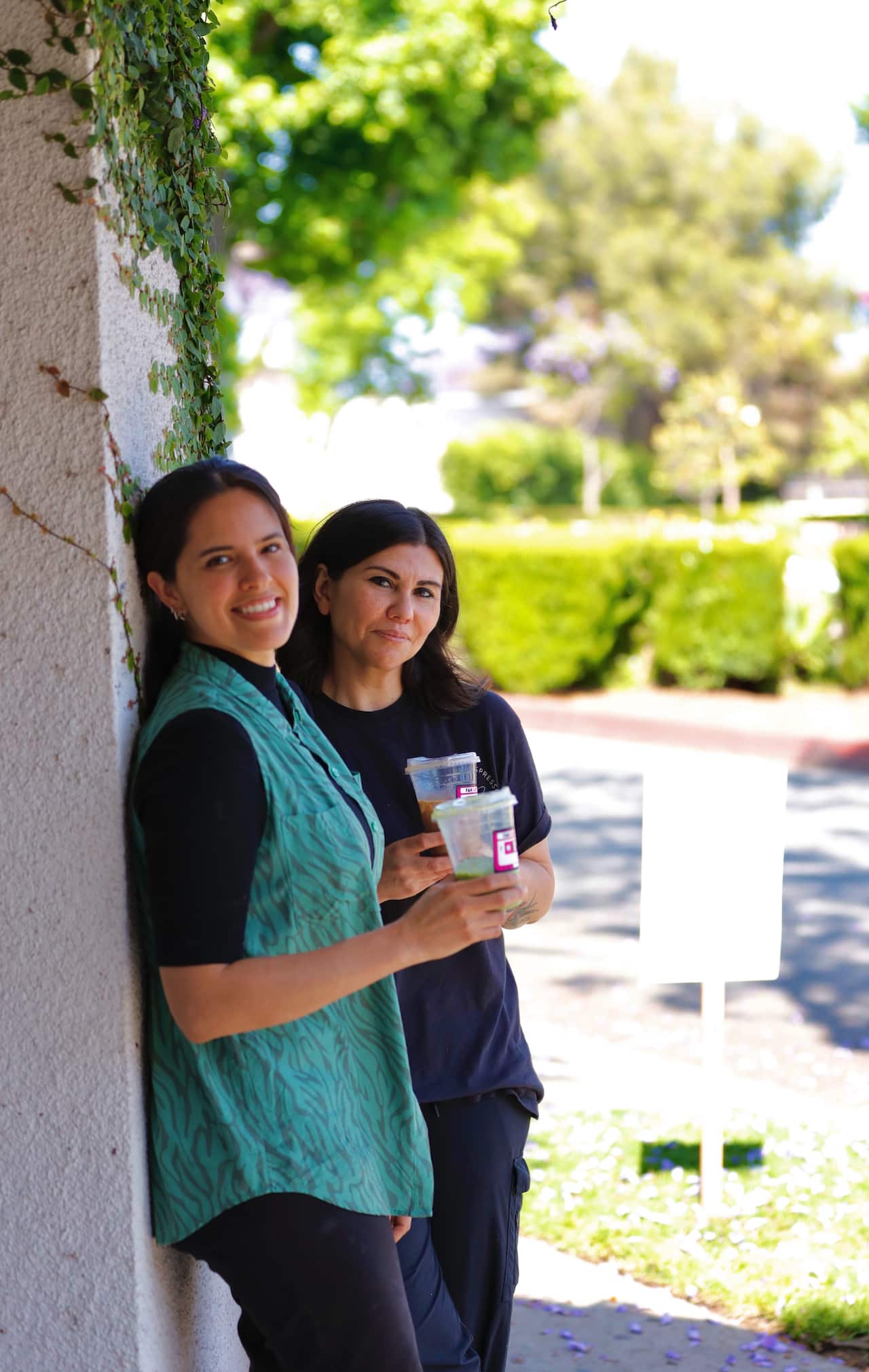 Two women holding plastic coffee cups stand next to a wall outside.
