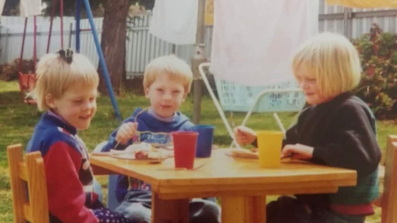 a vintage photo of three young blonde children sitting at a kids table in backyard with laundry hanging out