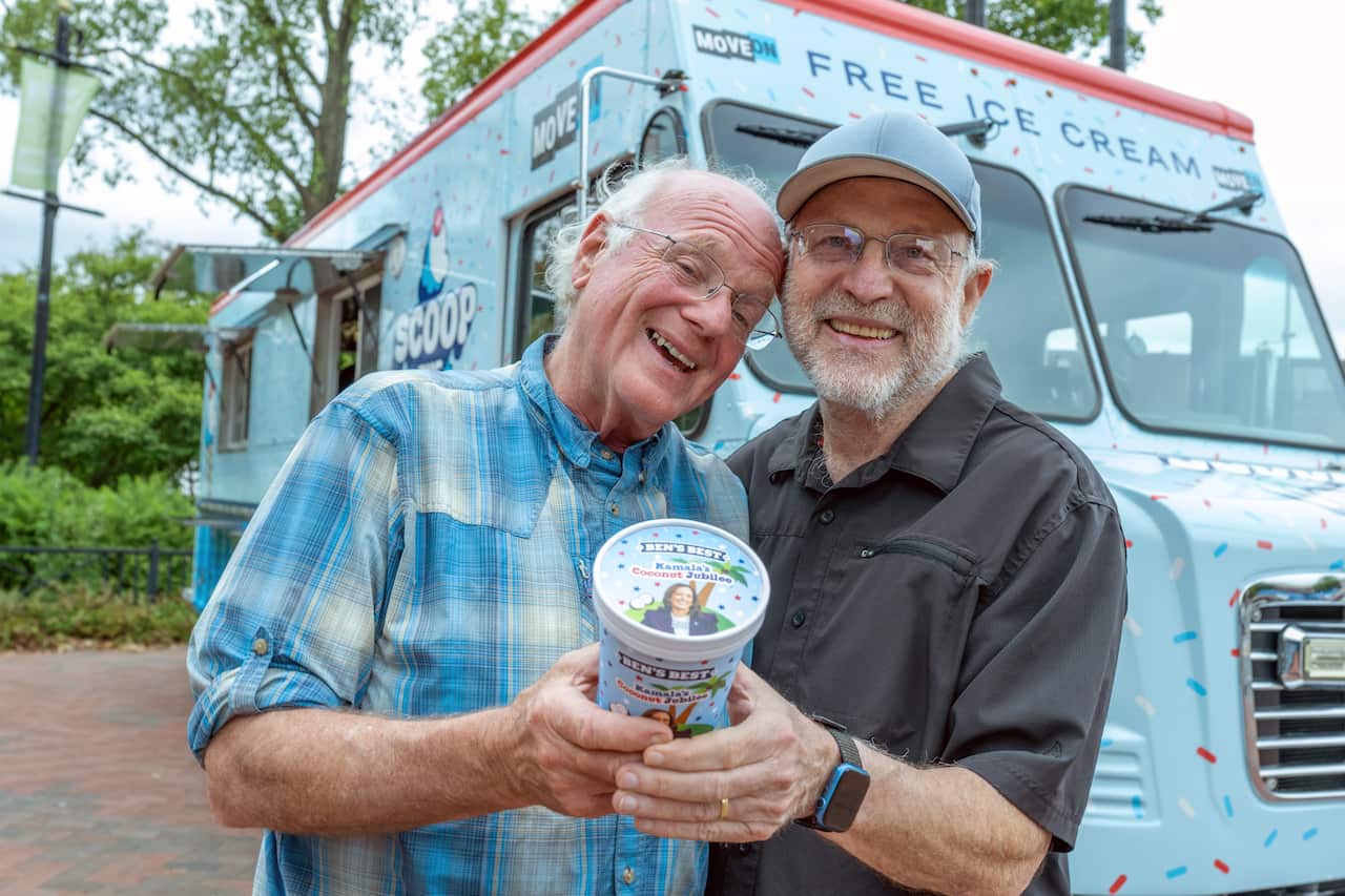 Two men holding a tub of ice-cream that has Kamala Harris' face on it. They're standing in front of a light blue van.