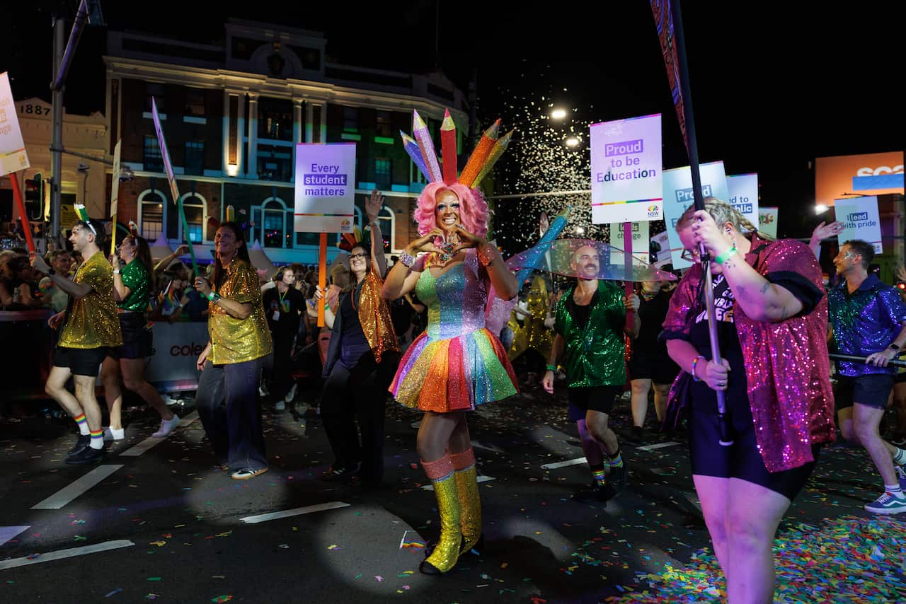 A vibrant night scene from a parade featuring a central performer in a sparkling, rainbow-colored dress and pink hair. They are wearing a unique headpiece designed to look like a fan of oversized, colorful pencils and are making a heart shape with their hands. Surrounding them are other participants wearing sequined shirts in gold, green, and pink, some holding signs that read "Every student matters" and "Proud to be Education". Confetti fills the air against the backdrop of city buildings and a crowd of spectators.