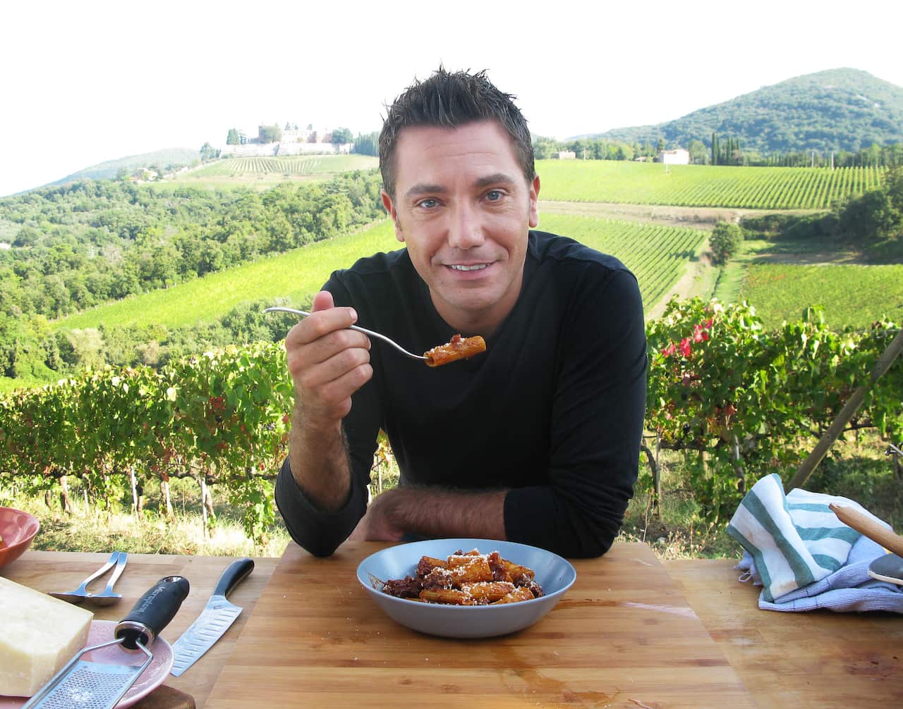 Gino eats a bowl of pasta with an Italian vineyard behind him
