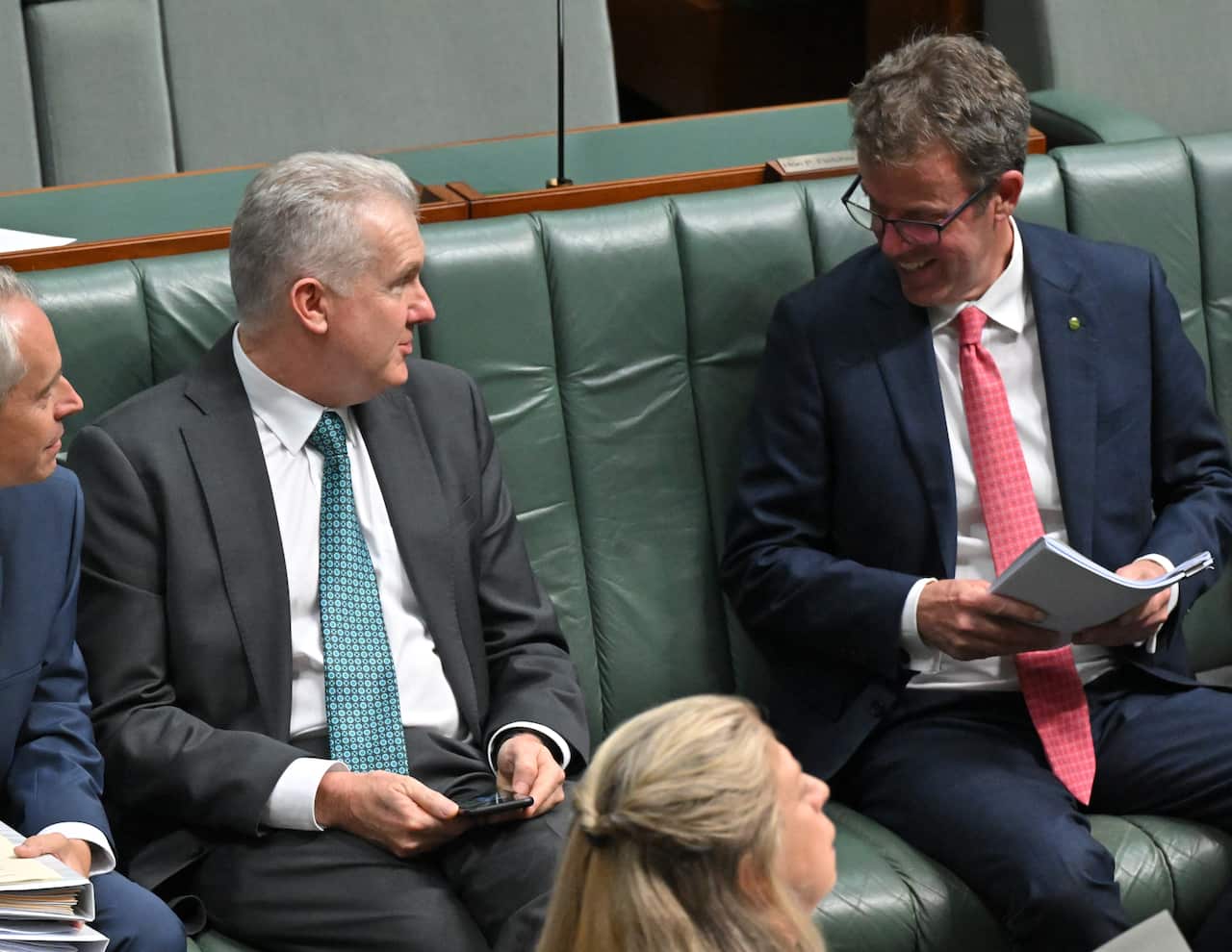 Two men sit on a green couch, facing each other as they talk.