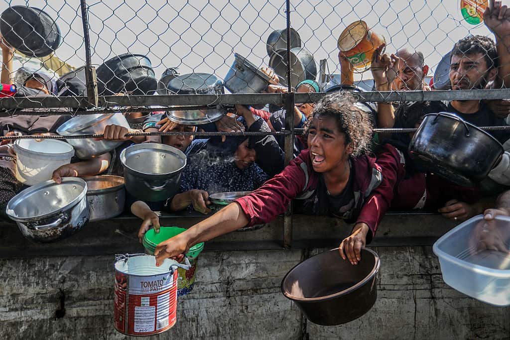 People stretch their arms through a metal fence with empty pots and pans, trying to receive food. 