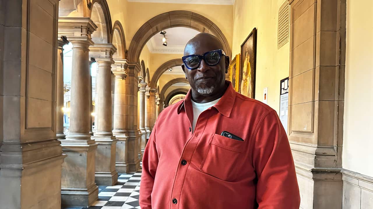 A middle-aged Black man wearing reading glasses, a red shirt and a white shirt underneath it stands in a museum hallway 