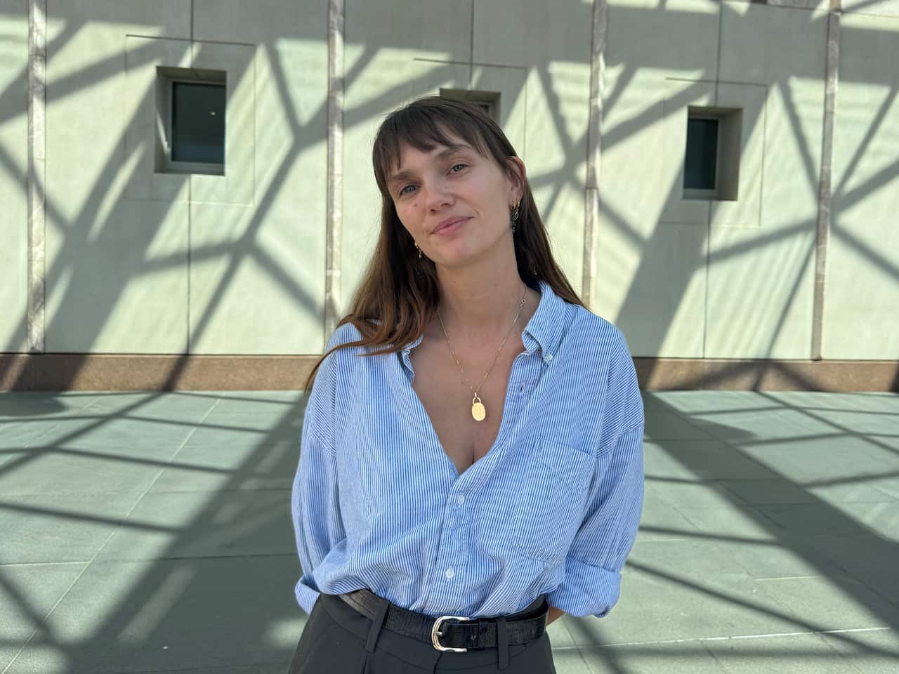 A young woman wearing a blue and white striped shirt and grey trousers with a black belt is smiling.