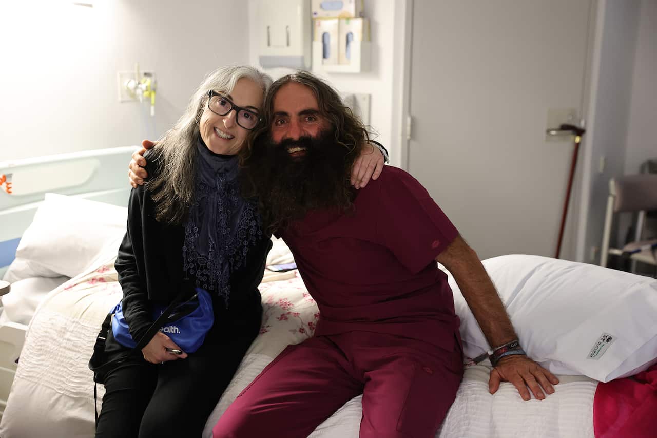 A woman and a man sit on a hospital bed, arms around each other's shoulders. He is wearing maroon scrubs. 