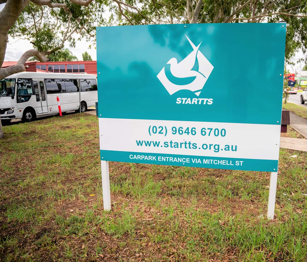 A green sign saying STARTTS stands on a grassy strip with a min-van in the background.