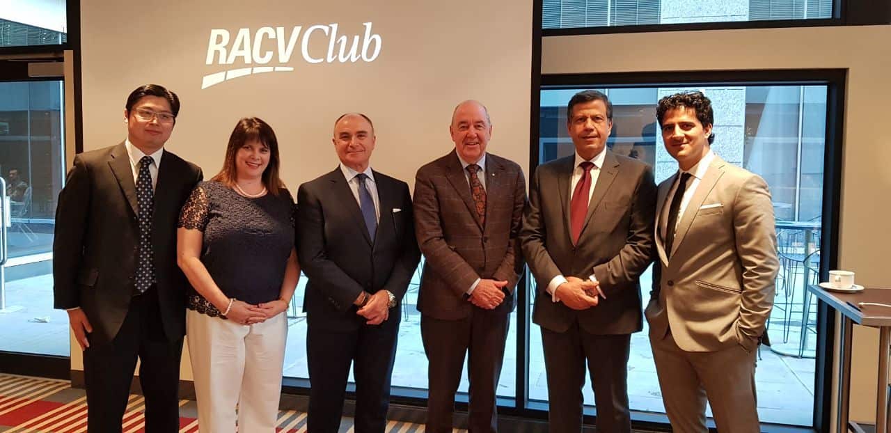 A group of six formally dressed people stand in a line in front of a screen displaying the "RACV Club" logo. They are posed for a group photo indoors, with large windows to the right showing an outdoor patio area.