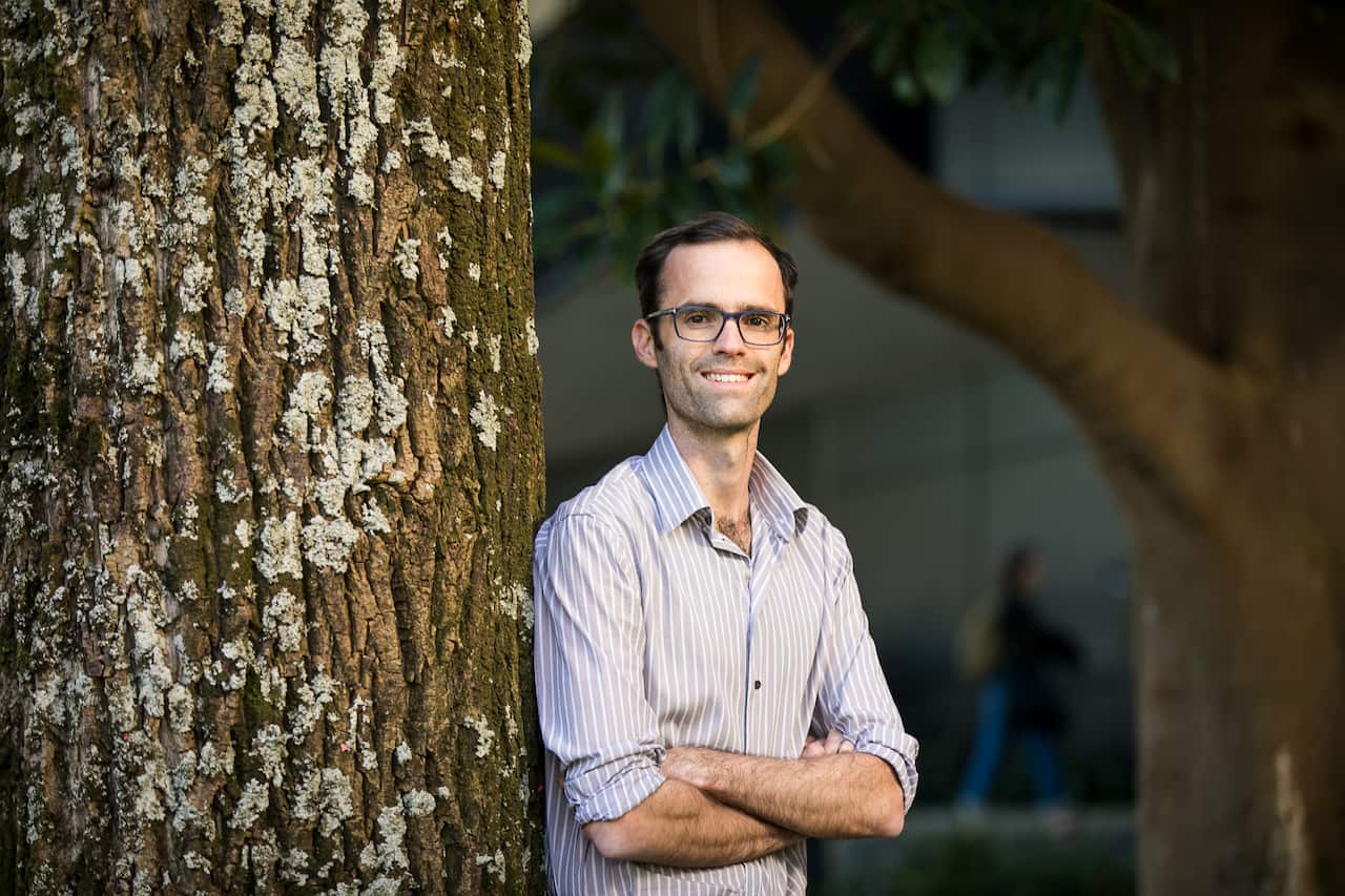 A man wearing glasses and a button-up shirt, with his arms folded leaning against a tree trunk.