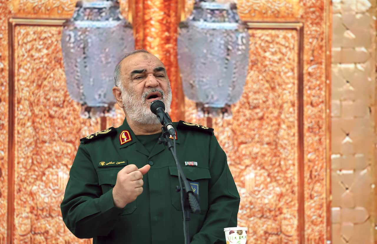A man in a green general's jacket holds his fist up while making a speech into a microphone in front of an orange backdrop