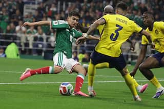 A footballer wearing a green shirt and white shorts attempts to pass or shoot the ball while being closely defended by multiple players.