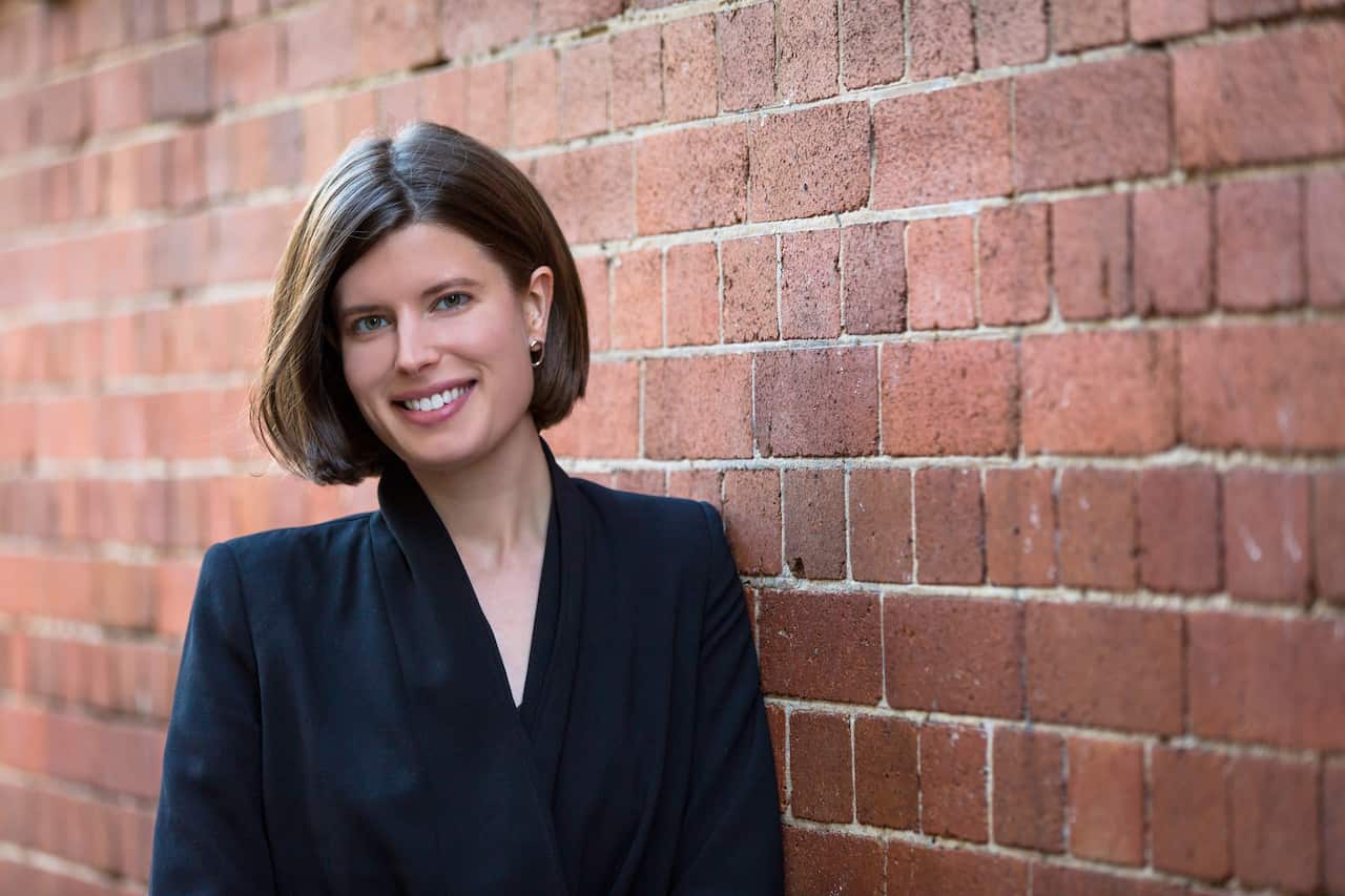 A woman with short hair wearing a suit. She's smiling against a red brick wall.