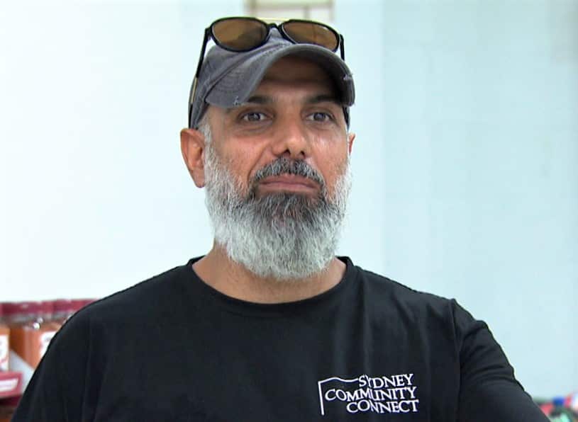 A man with a beard and wearing a black t-shirt stands facing camera.