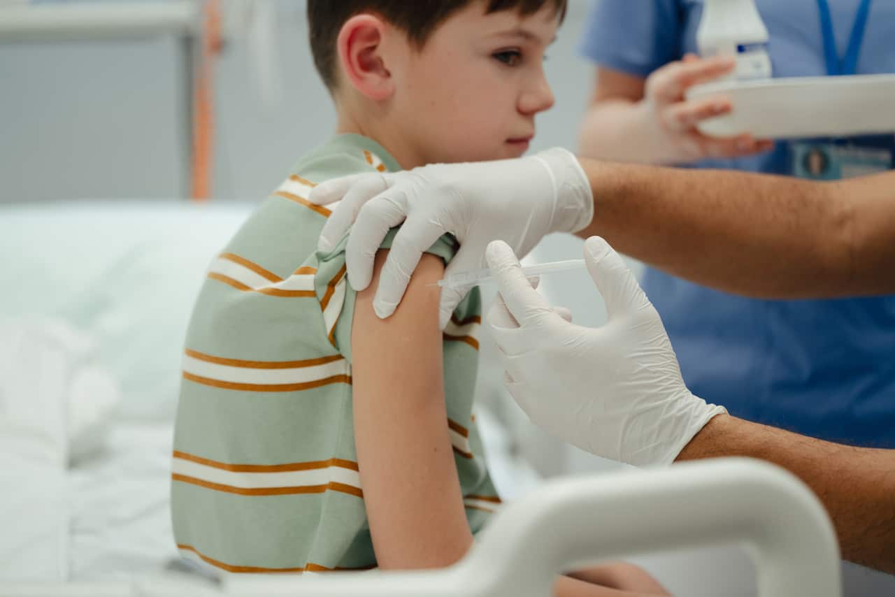 A young boy looks away with a neutral expression as a healthcare worker in white gloves administers a vaccination into his upper arm.