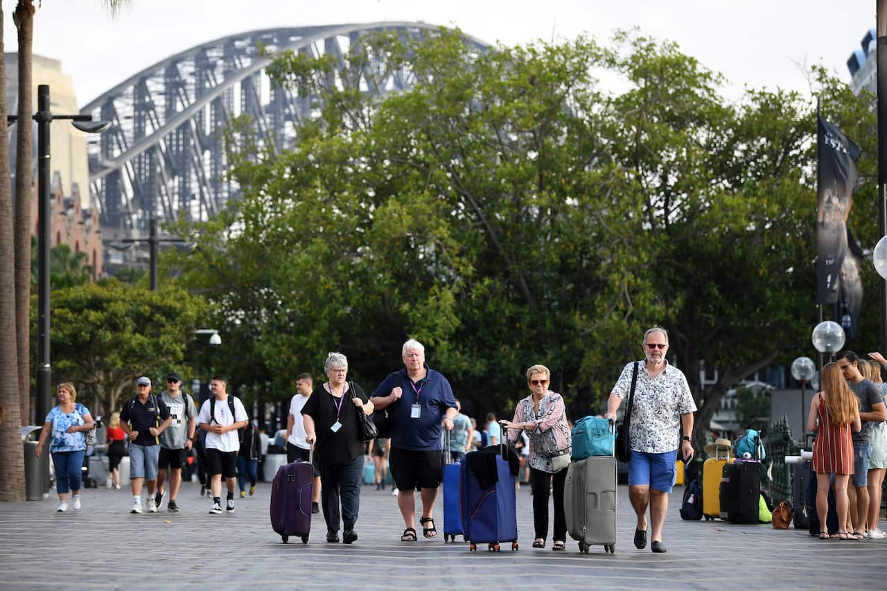 A crowd of people walk through the streets of Sydney. The Sydney Harbour Bridge is in the background, and tourists wheel suitcases in the foreground.