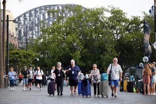 A crowd of people walk through the streets of Sydney. The Sydney Habour Bridge is in the background and tourists wheel suitcases in the centre of the crowd.