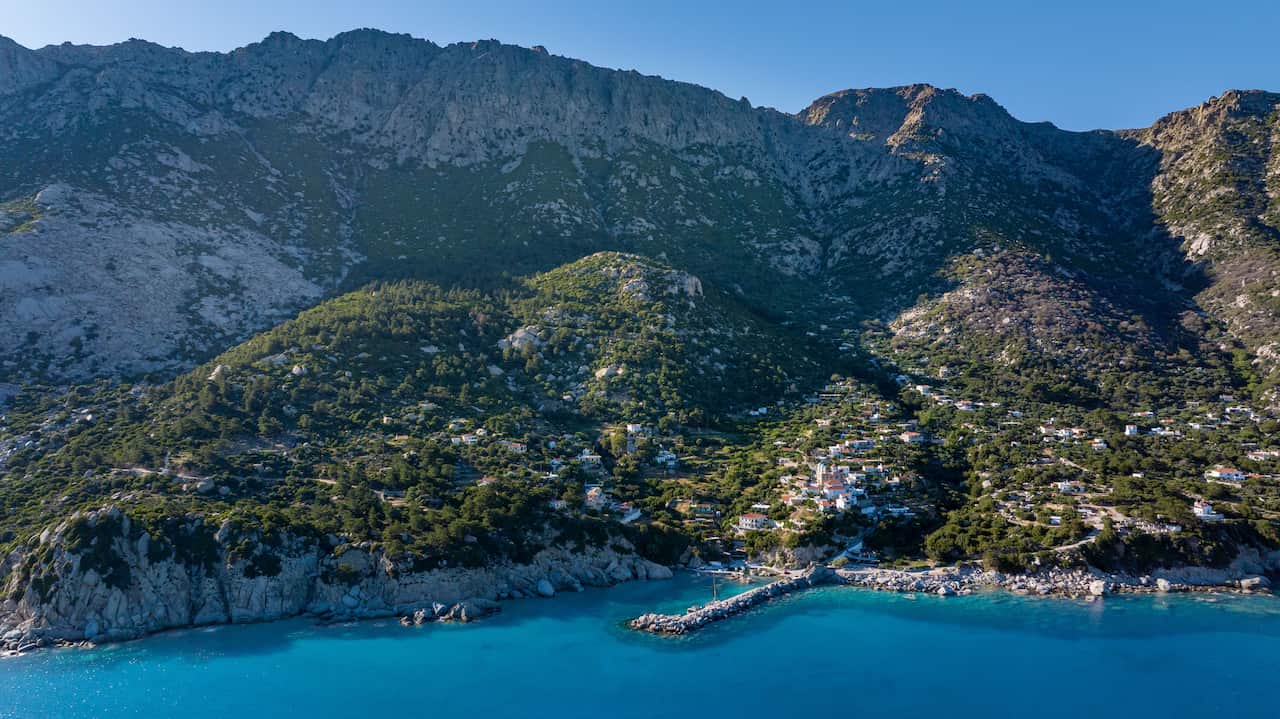 An island with rocky cliff faces emerges from a bright blue ocean. Large swathes of the island are covered in green forest, and clusters of white houses are visible. There is also a stone jetty.