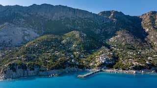An island with rocky cliff faces emerges from a bright blue ocean. Large swathes of the island are covered in green forest, and clusters of white houses are visible. There is also a stone jetty.